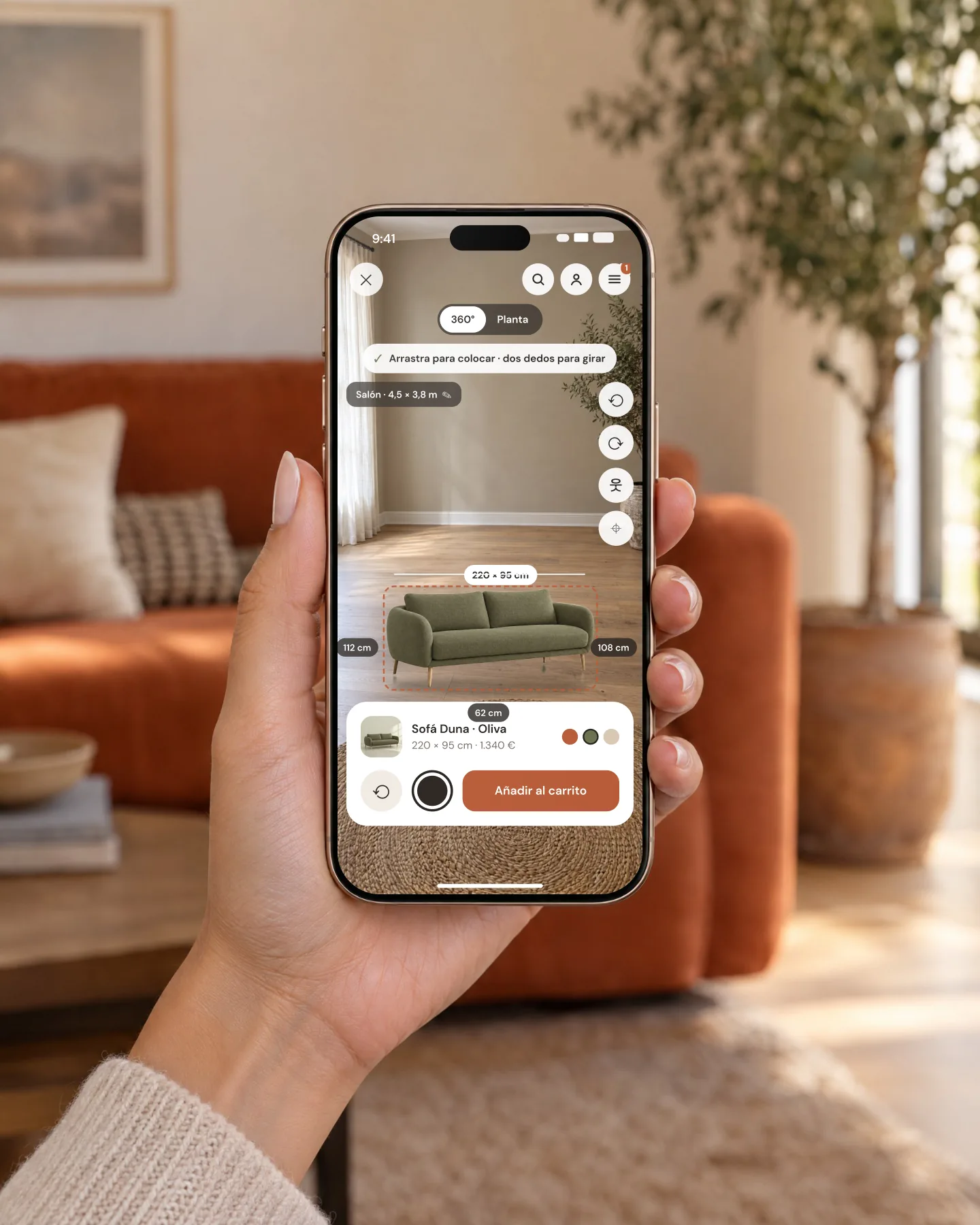 A hand holds an iPhone showing Olmo's AR viewer with the olive sofa placed and its measurements