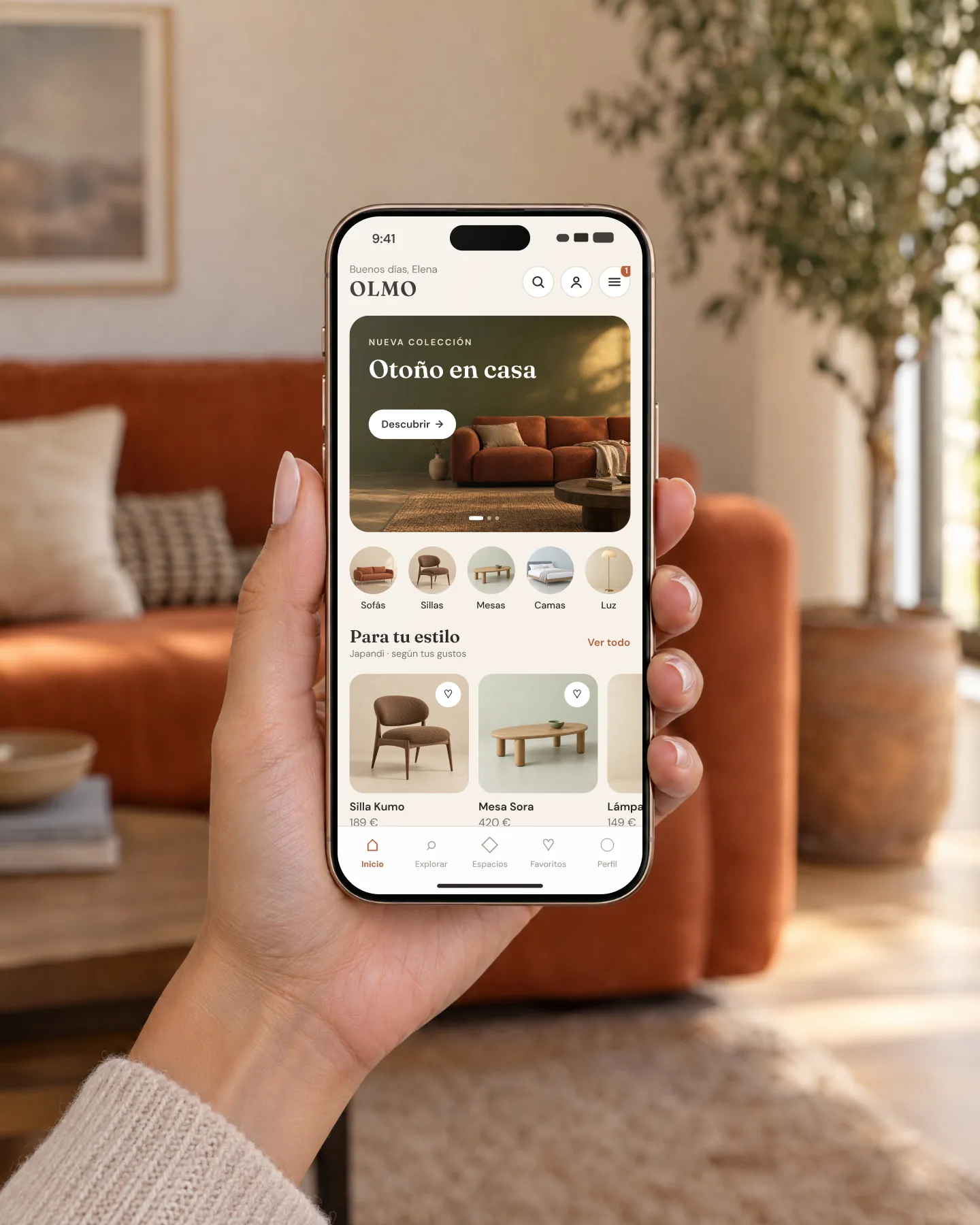 A hand holds an iPhone showing the Olmo home screen in a living room with a terracotta sofa
